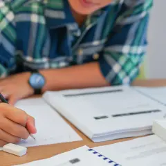 Uczeń pisze, zastanawiając się, czy przed "czy" stawiamy przecinek. Na biurku leżą książki i zeszyty.