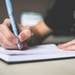 Dłoń trzymająca niebieski długopis, pisząca w notatniku. To stricte oznacza skupienie na zadaniu.