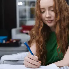 Ruda dziewczyna z burzą włosów pochyla się nad zeszytem, pisząc coś długopisem. W tle widać choinkę, co sugeruje, że to czas świąteczny, a jej skupienie przypomina mowę pozornie zależną – jakby była w swoim świecie.