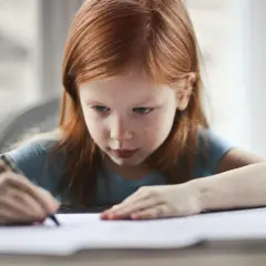 Ruda dziewczynka skupiona na rysowaniu. Jej dłoń trzyma ołówek, a wzrok skierowany jest na kartkę papieru.