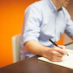 Mężczyzna pisze w notatniku, ćwicząc zasady interpunkcji. Obok laptop, na pomarańczowym tle.