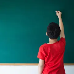 Chłopiec w czerwonej koszulce pisze kredą na zielonej tablicy, tworząc swoje pierwsze szkolne zadanie, oraz przygotowując się do lekcji.