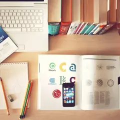 Biurko z laptopem, książkami i zeszytem. Kinestetyk tworzy szkice, inspirując się logotypami w otwartym albumie.
