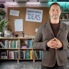 Mężczyzna w marynarce stoi przed tablicą z napisem "BRAINSTORMING IDEAS". W związku z tym, otoczenie sugeruje kreatywne środowisko pracy.