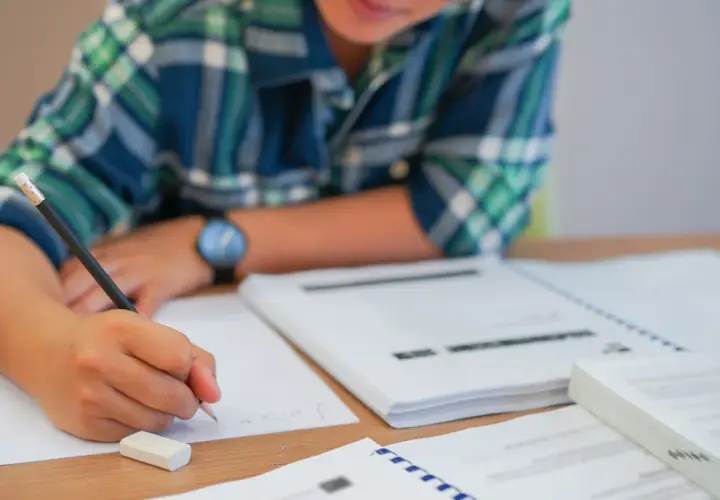Uczeń pisze, zastanawiając się, czy przed "czy" stawiamy przecinek. Na biurku leżą książki i zeszyty.