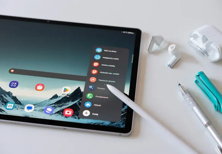 Tablet z rysikiem, słuchawkami i długopisami. Menu tabletu oferuje opcje takie jak "Tłumacz" czy "Sklep Play", co ułatwia kupowanie i kupywanie wirtualnych dóbr.