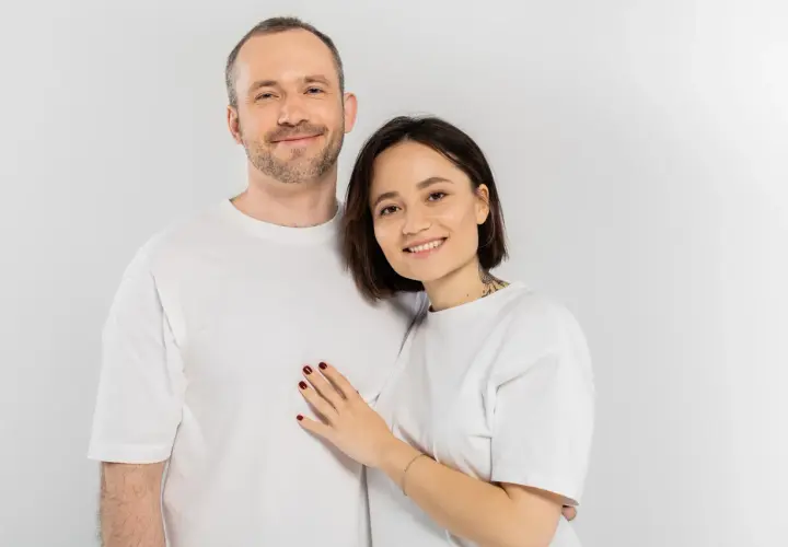 Uśmiechnięta para w białych koszulkach. Ona kładzie dłoń na jego piersi. Pewnie zastanawiają się nad śmiesznymi pytaniami.