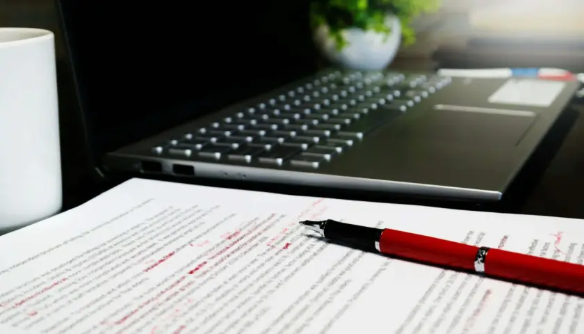 Czerwony długopis leży na dokumencie z poprawkami, obok laptopa. Widać tu różne odmiany cudzysłowu, które autor tekstu musiał poprawić.