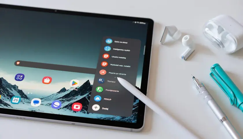 Tablet z rysikiem, słuchawkami i długopisami. Menu tabletu oferuje opcje takie jak "Tłumacz" czy "Sklep Play", co ułatwia kupowanie i kupywanie wirtualnych dóbr.
