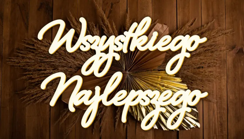 Napis "Wszystkiego najlepszego" ozdobiony suszonymi trawami i złotym wachlarzem na drewnianym tle.