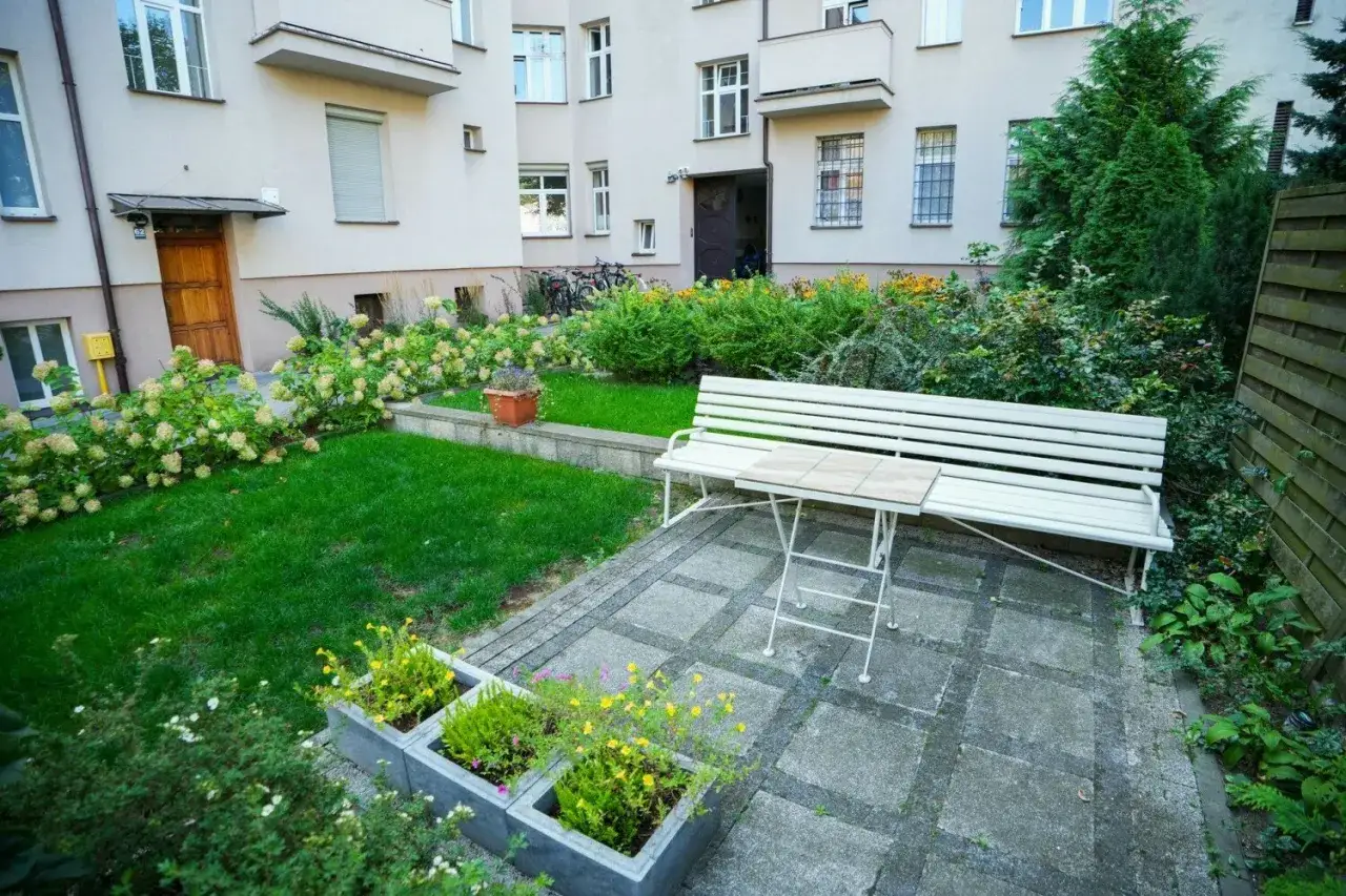 Urokliwe podwórze z ławką i stolikiem, otoczone zielenią i kwitnącymi krzewami. Idealne miejsce na chwilę relaksu.