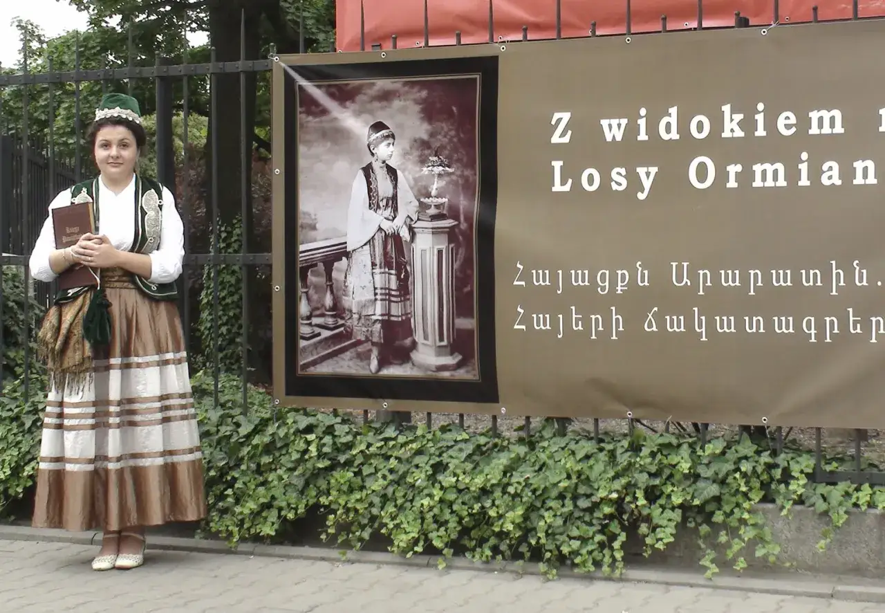 Dziewczyna w stroju ormiańskim trzyma książkę obok baneru